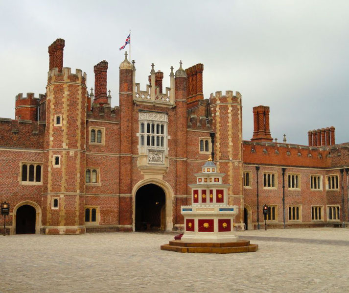 A historic red-brick palace with ornate chimneys, arched entryways, and a central decorated gateway. A colorful, tiered fountain stands in the cobbled courtyard, with a Union Jack flag flying above.