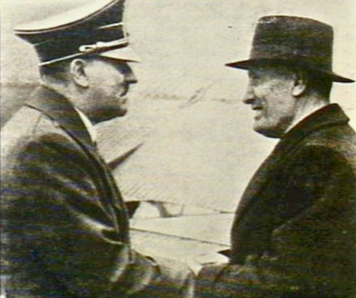 Two men in coats and hats shake hands in a historical black-and-white photo, suggesting a formal or diplomatic meeting.