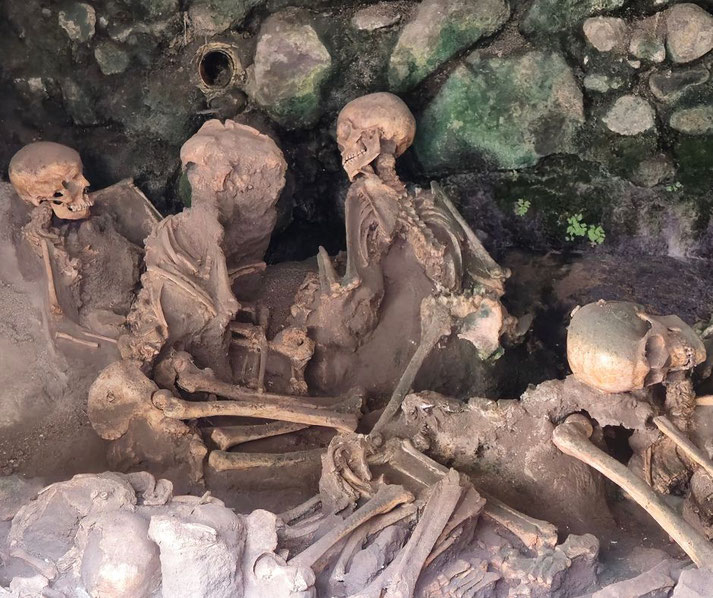 Several human skeletons lie in close proximity, partially preserved in volcanic ash, inside a stone-walled chamber with traces of moss and moisture.