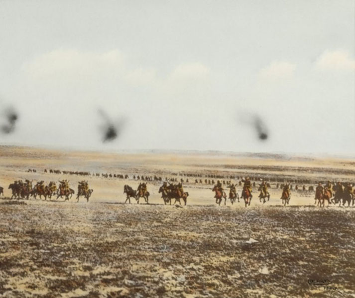 Mounted troops charge across a vast desert plain in formation, kicking up dust as distant cavalry lines stretch across the horizon under a pale sky.