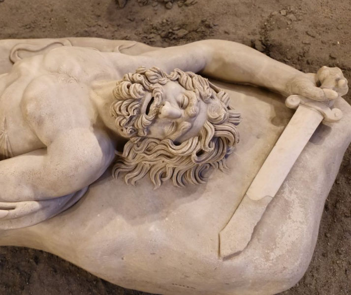 Marble statue of a fallen bearded warrior lying on a stone cushion, eyes closed, gripping a short sword, with a pained yet serene expression.