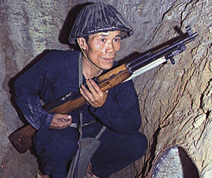A Viet Cong soldier crouches in a narrow tunnel, gripping a rifle with a bayonet, wearing dark clothing and a pith helmet.