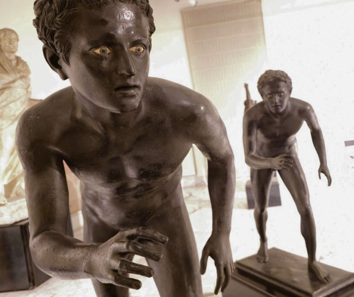 Bronze statues of two nude young runners in mid-motion, with intense expressions and inlaid eyes, capturing dynamic athleticism and focus.