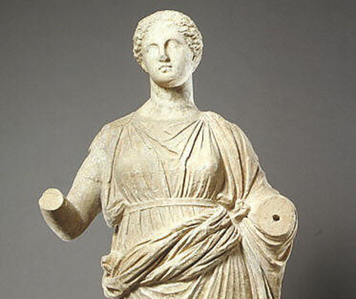 Ancient marble statue of a standing woman in a flowing gown with both arms missing, posed in a frontal stance.