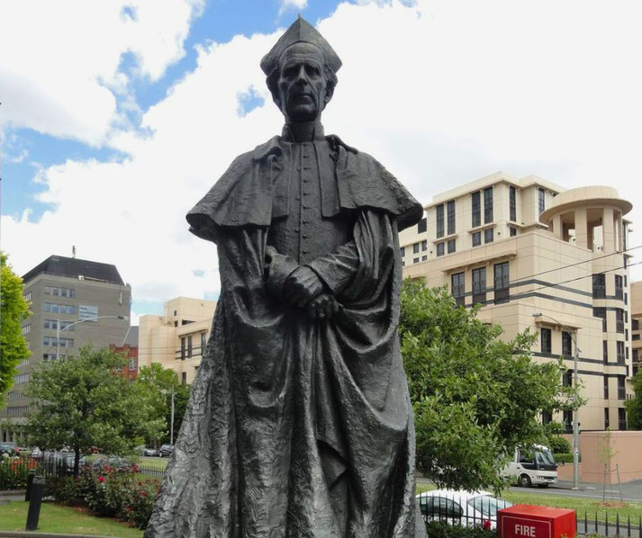 A statue of a robed religious figure wearing a bishop’s hat stands with hands clasped, set in an urban area with modern buildings and trees in the background.