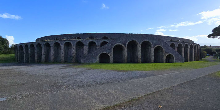 The amphitheatre of Pompeii, an ancient oval structure with multiple stone arches and grassy surroundings, set against a bright blue sky with scattered clouds.