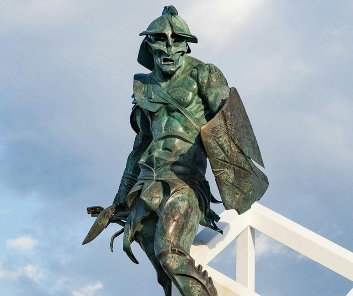A bronze statue of a warrior in an aggressive stance, wearing a crested helmet, armor, and wielding a sword and shield.