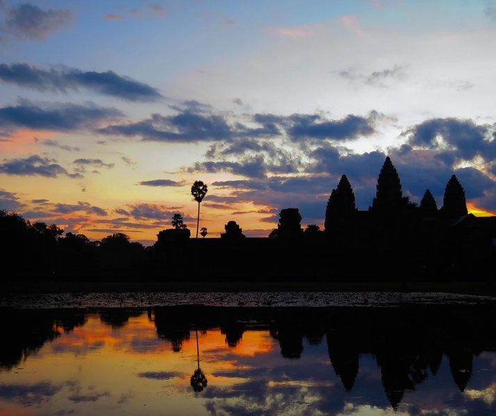 Silhouetted Angkor Wat temple and palm trees reflected in water during a vibrant sunset with colorful clouds.