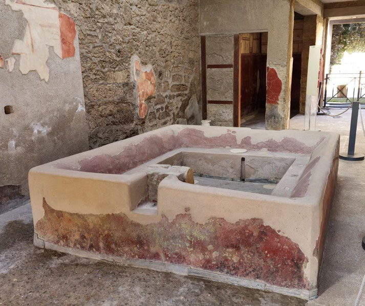 A large vat inside the Fullery of Stephanus in Pompeii, used for textile processing. The structure features a rectangular design, red and white plaster, and is surrounded by ancient stone walls.