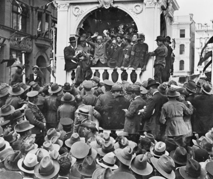 A large crowd in hats gathers around a balcony where men, some in uniform, stand addressing them. The scene appears historic, with festive banners and old-style architecture in the background.