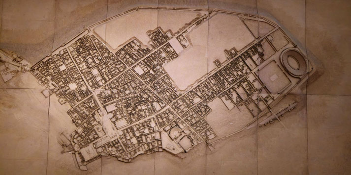 Detailed ancient city map showing a dense street grid, buildings, a large amphitheater on the right, and a fortified wall enclosing the settlement.