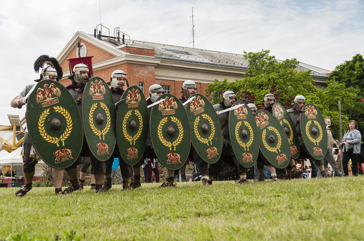 Roman reenactors in chainmail and helmets advance in formation with green oval shields adorned with laurel wreaths and eagle motifs.