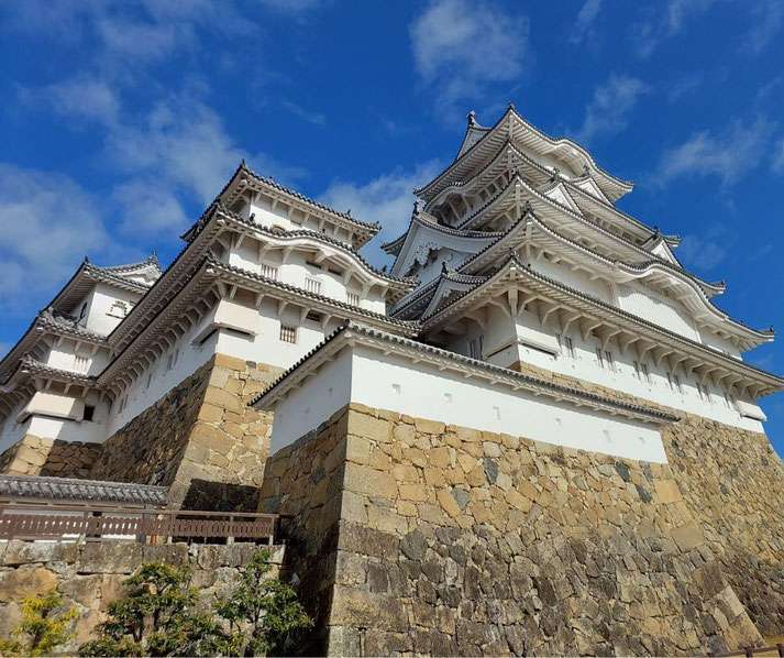 Himeji Castle, a stunning Japanese fortress with white walls, intricate wooden eaves, and a massive stone foundation, standing against a bright blue sky with scattered clouds.