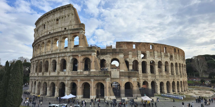 The Colosseum in Rome, a massive ancient amphitheater with tiered arches and partially ruined walls, surrounded by visitors and a cloudy sky.