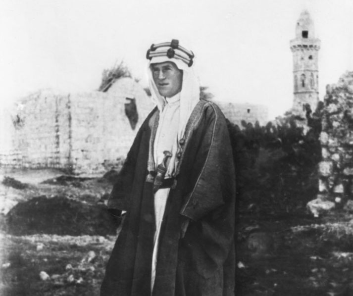 A man in traditional Arab attire stands in front of stone ruins and a tall tower, suggesting a historical Middle Eastern setting.