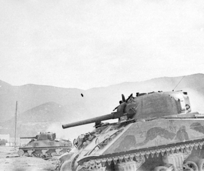 Two tanks in motion during combat, one firing a shell with a visible projectile in midair against a mountainous backdrop.