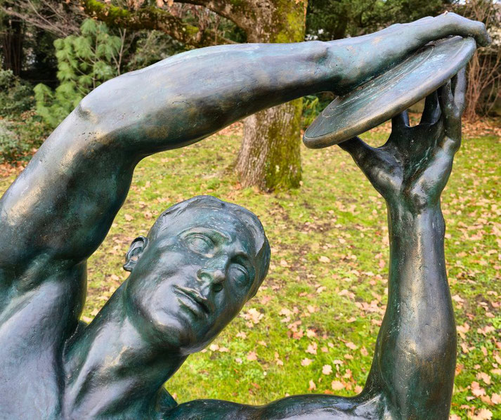 A bronze statue of a discus thrower in mid-motion, arms raised gracefully. The detailed sculpture is set against a background of grass, fallen leaves, and trees in a serene outdoor setting.