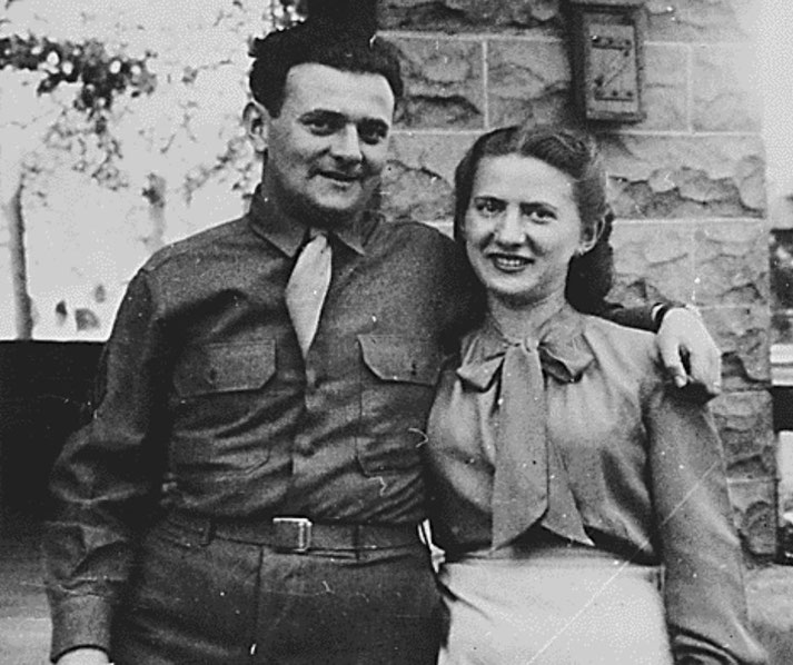 Black and white photo of a smiling couple; the man wears a military uniform and the woman is in a blouse with a bow, standing close together in front of a stone building.
