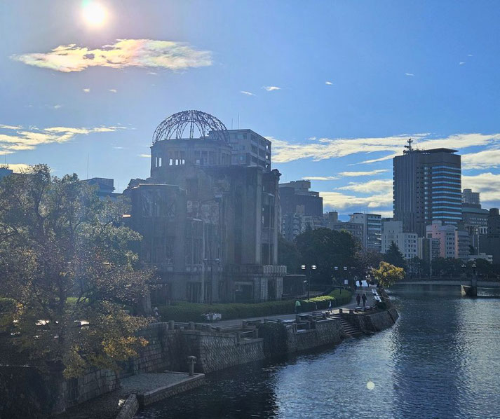 The Atomic Bomb Dome stands by the river under bright sunlight, surrounded by trees and city buildings in modern Hiroshima.