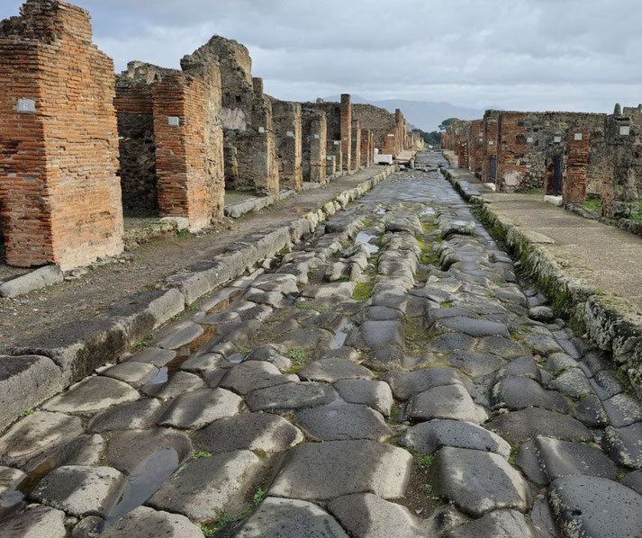 A cobblestone street in Pompeii, with grooves worn by ancient carts, surrounded by brick ruins of buildings, and a cloudy sky in the background.