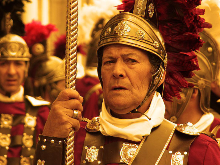 Man in ornate Roman-style armor and helmet with red plumes, holding a staff, among others in similar ceremonial attire.