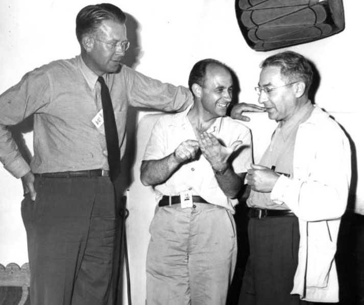 Three men engaged in lively conversation, one gesturing animatedly while the others listen and smile in a casual indoor setting.