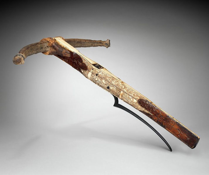 A historical crossbow with intricate carvings on its wooden body. The crossbow is mounted on a stand, and its string is partially visible.