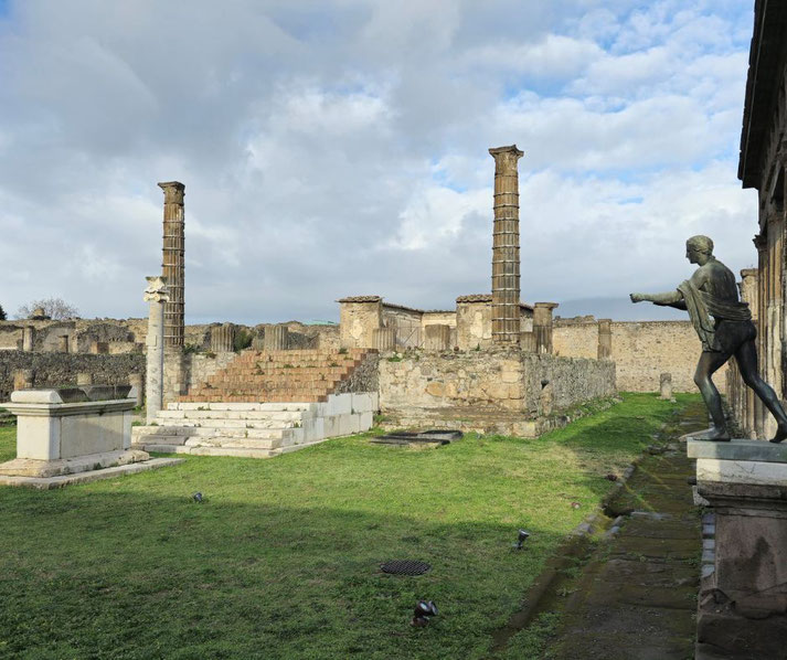 The Temple of Apollo at Pompeii, featuring ancient columns, a statue of Apollo, and a stone altar amidst ruins, with a grassy courtyard and a cloudy sky in the background.