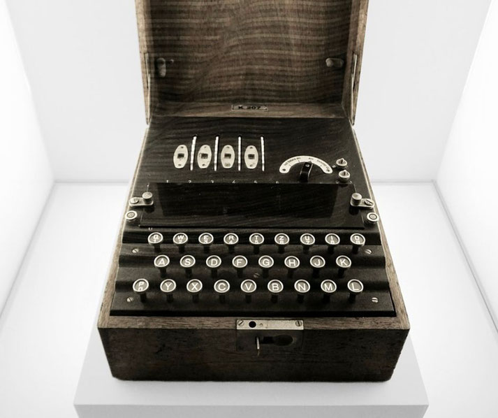A vintage Enigma machine with a wooden case and circular keys is displayed in a white exhibition space.