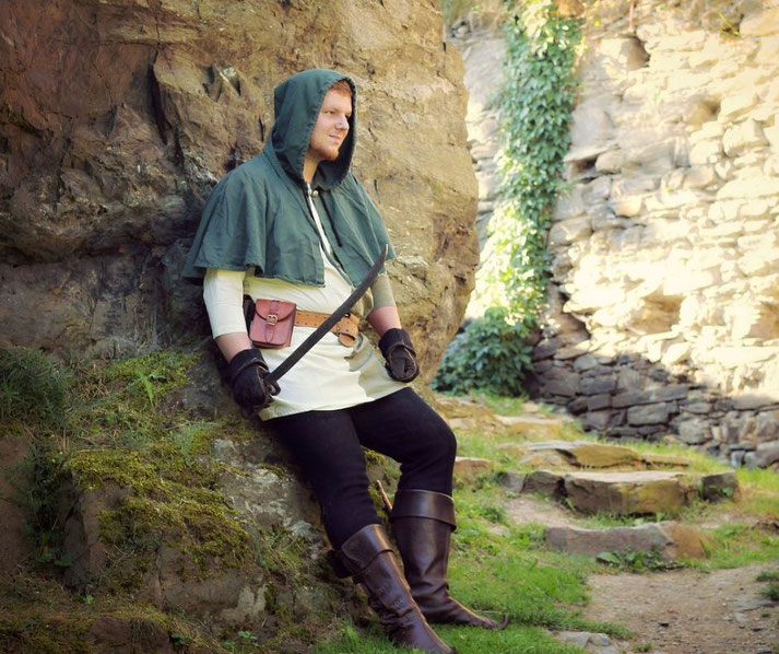 A man dressed as a medieval rogue or ranger leans against a rock, wearing a green hooded cloak, tunic, gloves, and boots.