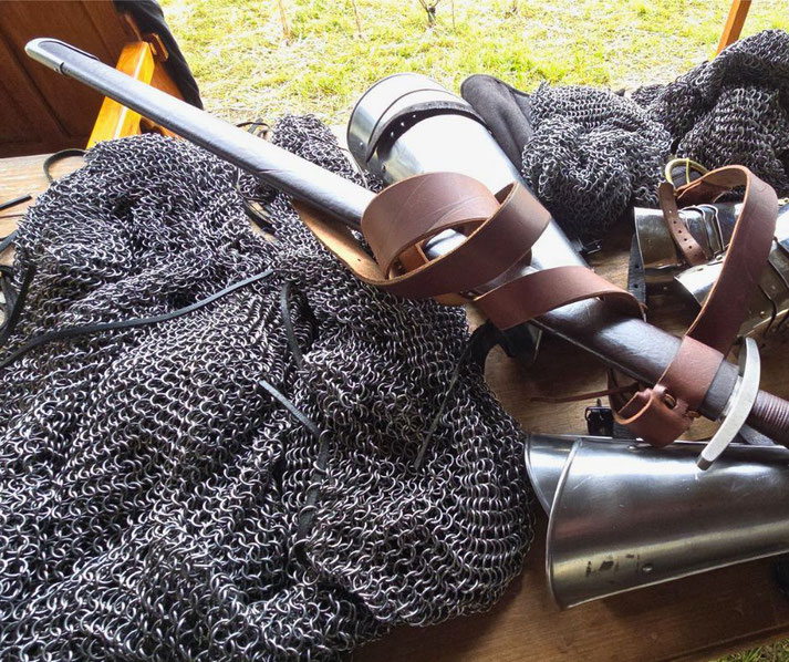 A medieval reenactment setup with chainmail, a longsword wrapped in leather straps, and pieces of steel armor, including gauntlets and greaves, laid out on a wooden table outdoors.
