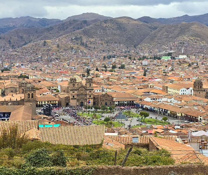 A panoramic view of Cusco, Peru, showcases the historic Plaza de Armas, colonial cathedrals, and terracotta rooftops, with the Andean mountains and "El Perú" inscription in the background.