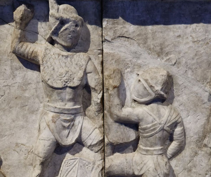 Stone relief of two gladiators in combat, both wearing helmets, arm guards, and short tunics, with raised arms in dynamic fighting poses.