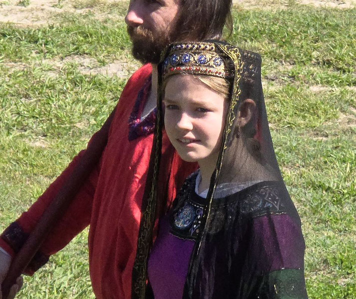 A young girl in a medieval-style gown and ornate headpiece with a black veil stands beside a bearded man in a red tunic.