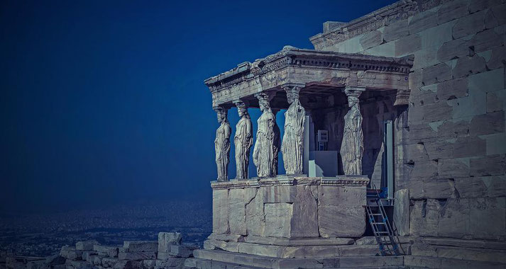 Ancient Greek temple porch featuring six sculpted female figures as columns, set against a deep blue sky on an elevated stone platform.