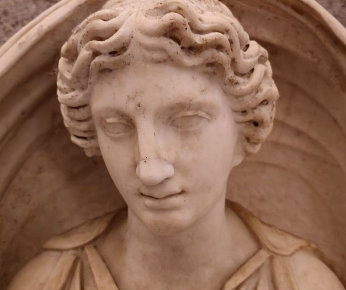 Close-up of a marble statue's face with soft features, wavy hair, and a faint smile, framed by a rounded architectural background.