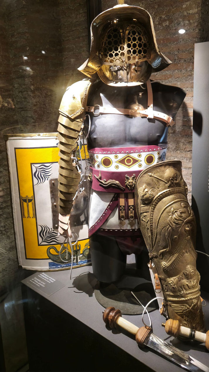 Gladiator armor display featuring a metal helmet with honeycomb visor, arm and leg guards, decorative belt, sword and shield in museum case.