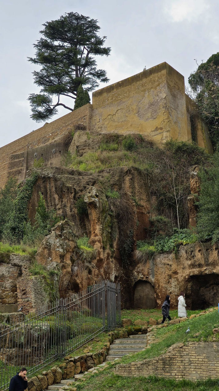 Ancient rocky hillside with arched cave entrances, topped by weathered yellow walls and a tall tree, with visitors walking below on stone steps and grassy paths.