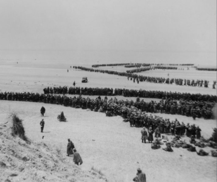 Thousands of soldiers stand in long lines on a beach, waiting for evacuation, with the sea in the background and minimal equipment visible.