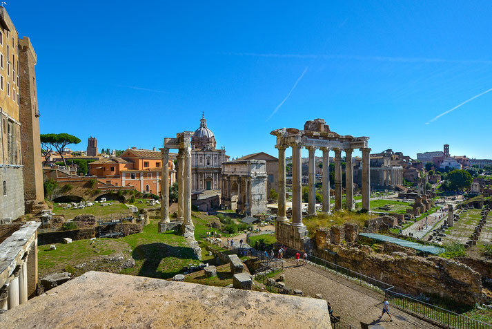 Daytime view of the Roman Forum showing ancient ruins, standing columns, and surrounding Renaissance and Baroque buildings.