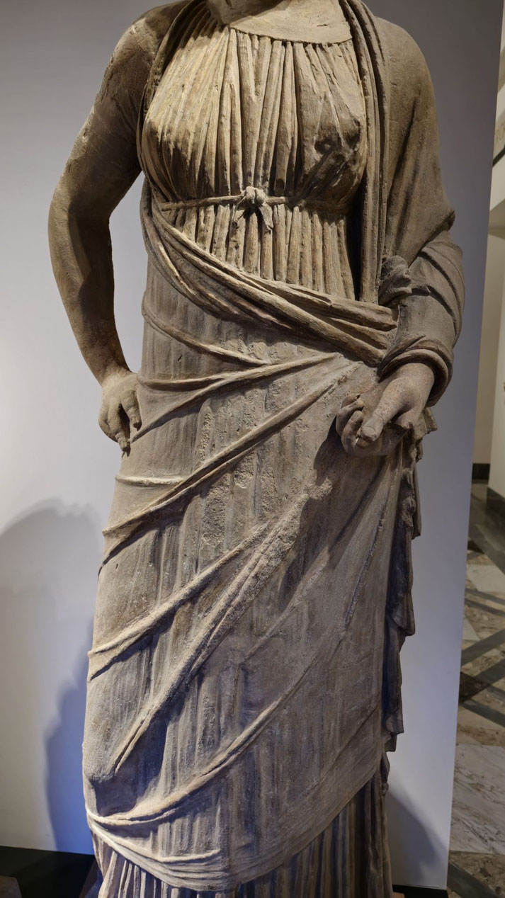 Ancient stone statue of a woman in a draped, pleated garment with one arm bent at the waist and the other holding fabric.