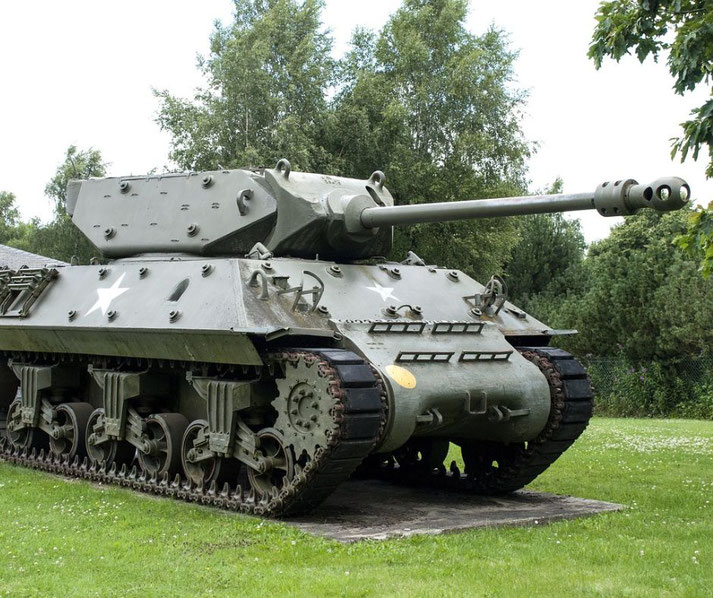 A green WWII-era tank with a long barrel sits on display in a grassy park surrounded by trees.