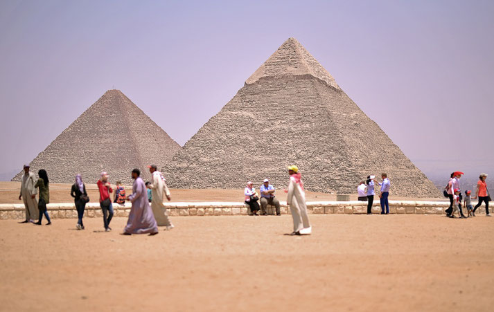 Pyramids of Giza in Egypt, with visitors walking and resting nearby. The iconic structures rise majestically from the desert under a clear sky, symbolizing ancient architectural brilliance.
