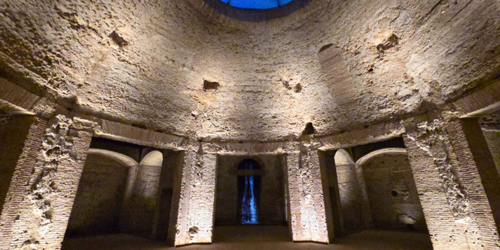 Circular ancient brick chamber with high domed ceiling, arched niches, and dramatic lighting highlighting its rough texture and architectural symmetry.