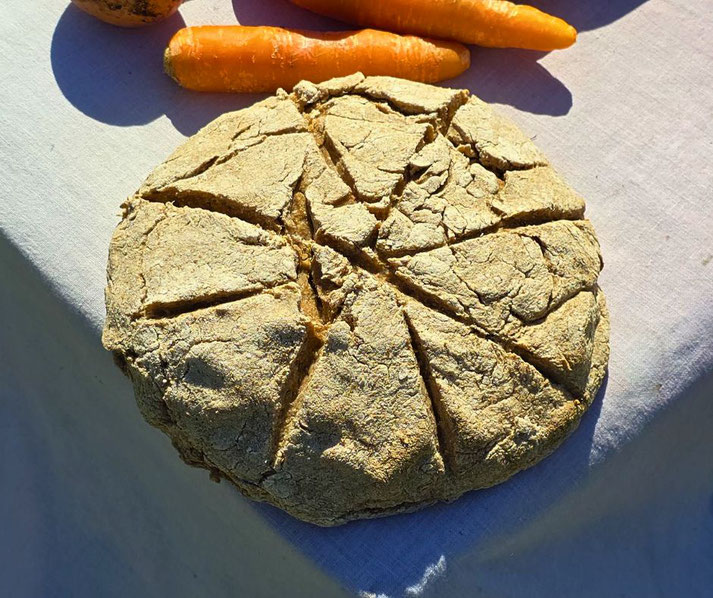 Rustic round loaf of bread with deep scoring lines, resting on a white cloth beside orange carrots in sunlight.