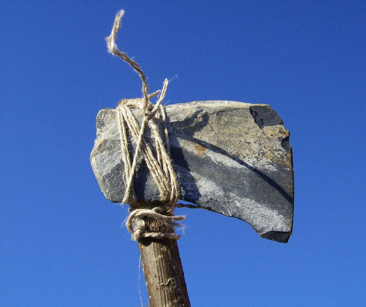 A prehistoric stone axe with a sharp-edged rock tied to a wooden handle using twine. The tool, set against a clear blue sky, resembles early human craftsmanship for cutting or hunting.