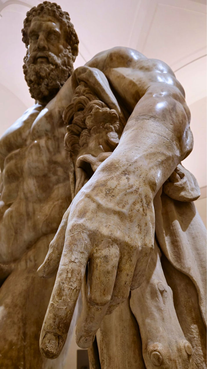Low-angle view of a powerful bearded statue with outstretched hand, holding a lion pelt—depicting strength and heroism, likely representing Hercules.