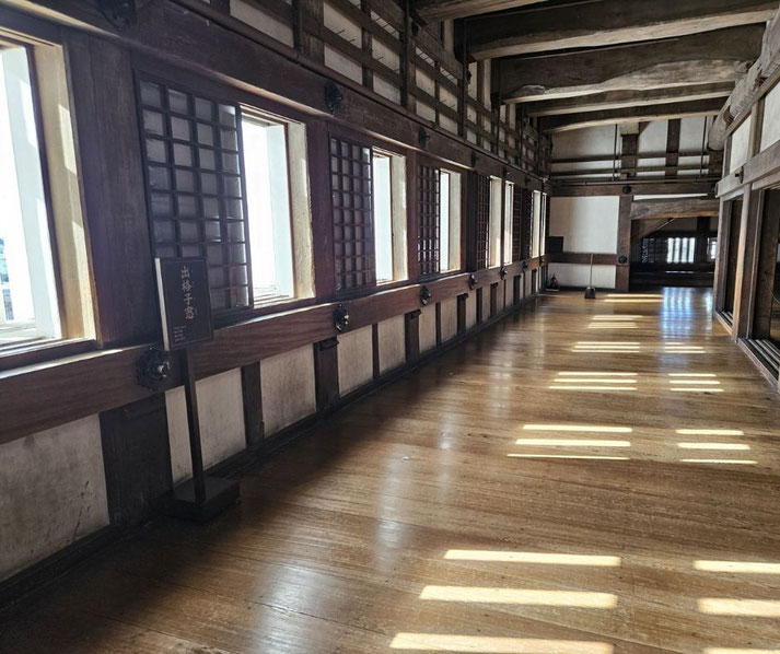 The interior of Himeji Castle, featuring a wooden corridor with latticed windows, exposed beams, and polished floors. Sunlight streams in, casting rectangular patterns on the floor.