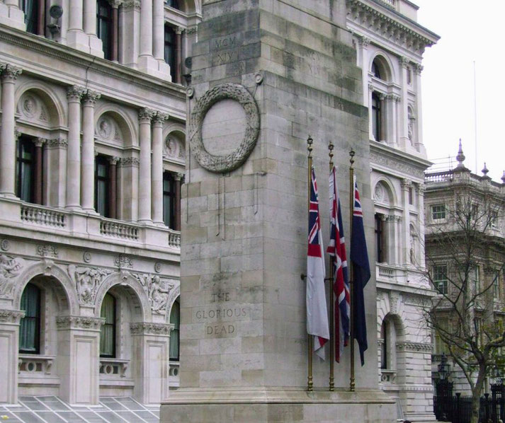 Stone war memorial with engraved wreath and the words “The Glorious Dead,” flanked by several British flags, set in front of a historic building.