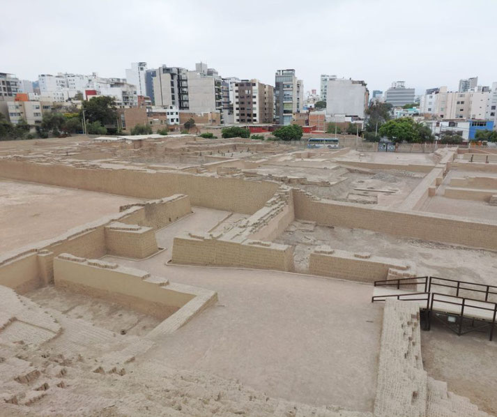 Huaca Pucllana, an ancient adobe pyramid in Lima, Peru. The archaeological site features clay walls and structures, contrasting with the modern city buildings in the background.